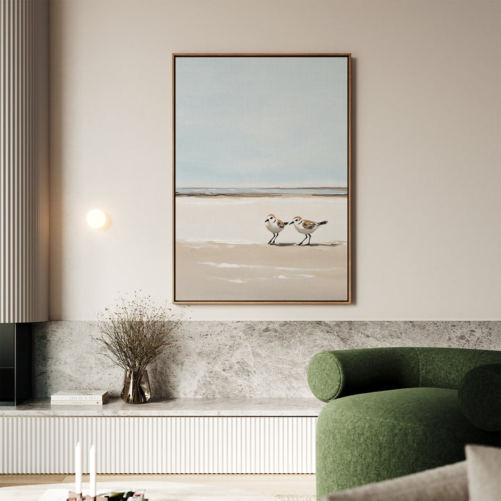 A minimalist living room featuring a green sofa, wall-mounted light, and the Coastal Chase Canvas Art depicting two shorebirds on sand, hanging above a marble shelf with dried flowers and stacked books.