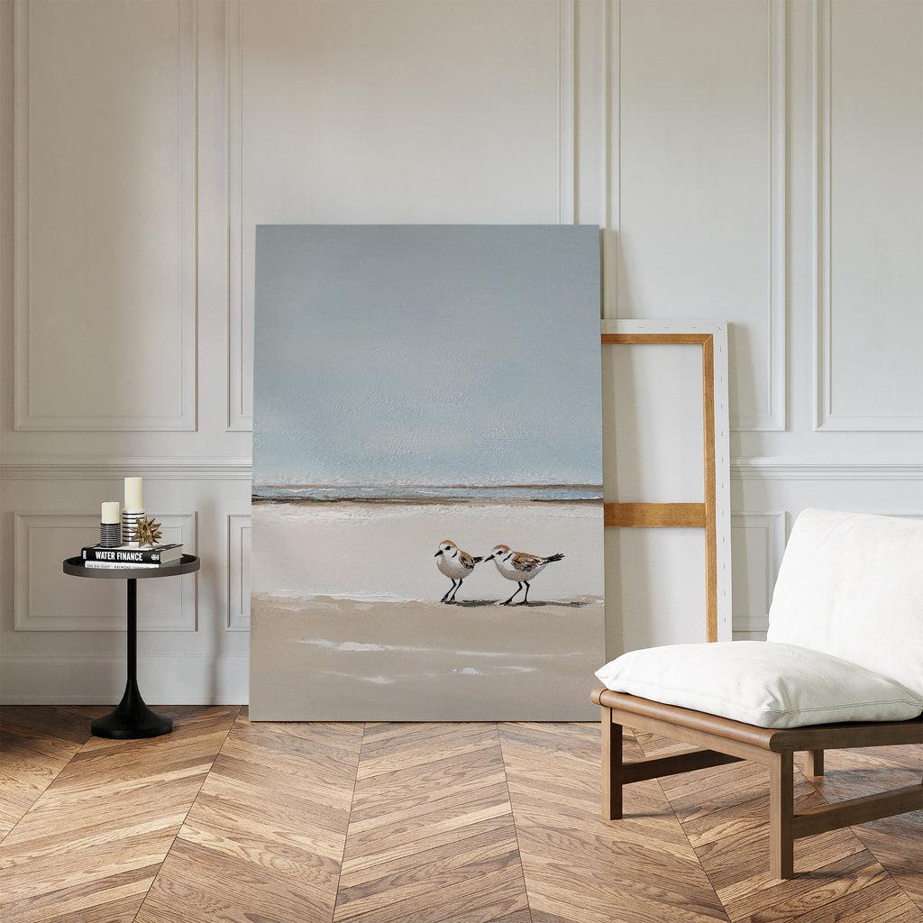 The Coastal Chase Canvas Art, featuring two shorebirds on a beach, leans against a white paneled wall beside an empty frame, modern black side table with candles, and cushioned chair on a herringbone wood floor.