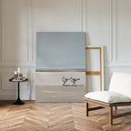 The Coastal Chase Canvas Art, featuring two shorebirds on a beach, leans against a white paneled wall beside an empty frame, modern black side table with candles, and cushioned chair on a herringbone wood floor.