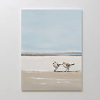 The Coastal Chase Canvas Art features two shorebirds on a sandy beach with calm water and a soft blue sky, creating serene wall décor perfect for any space.