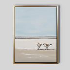 The Coastal Chase Canvas Art features two small shorebirds on a sandy beach with calm waves and a pale blue sky. The gold frame adds an elegant touch to this minimalist, serene wall art print.