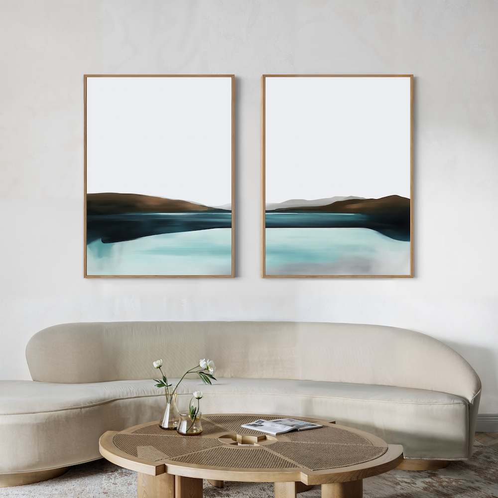 A modern living room features a curved beige sofa, a round wooden coffee table with vases and flowers, and the Coastal Calm Set of 2 abstract landscape paintings in blue, black, and brown tones on a light wall for a serene ambiance.