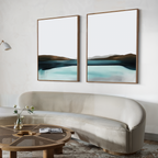 A modern living room features a curved beige sofa, a round wooden coffee table with a book and vase, and the Coastal Calm Set of 2 abstract landscape artworks in blue and brown tones on a light wall—ideal for contemporary interiors.