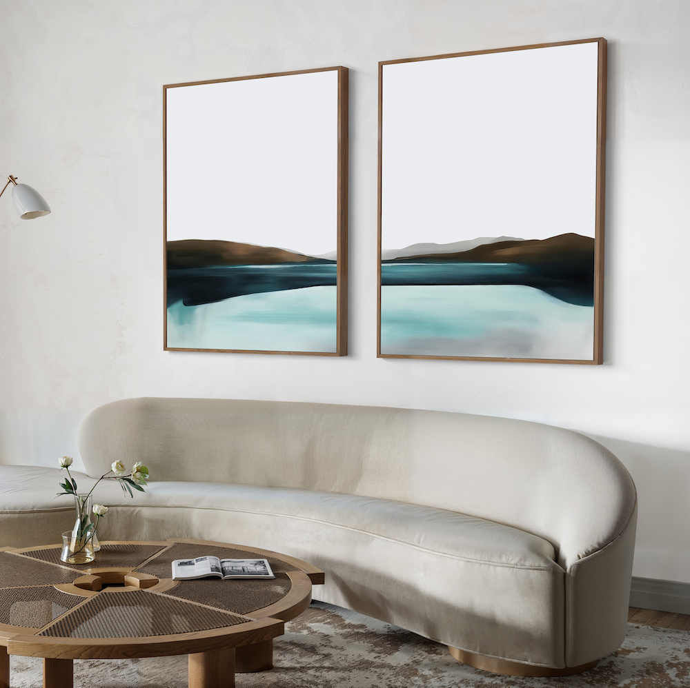 A modern living room features a curved beige sofa, a round wooden coffee table with a book and vase, and the Coastal Calm Set of 2 abstract landscape artworks in blue and brown tones on a light wall—ideal for contemporary interiors.