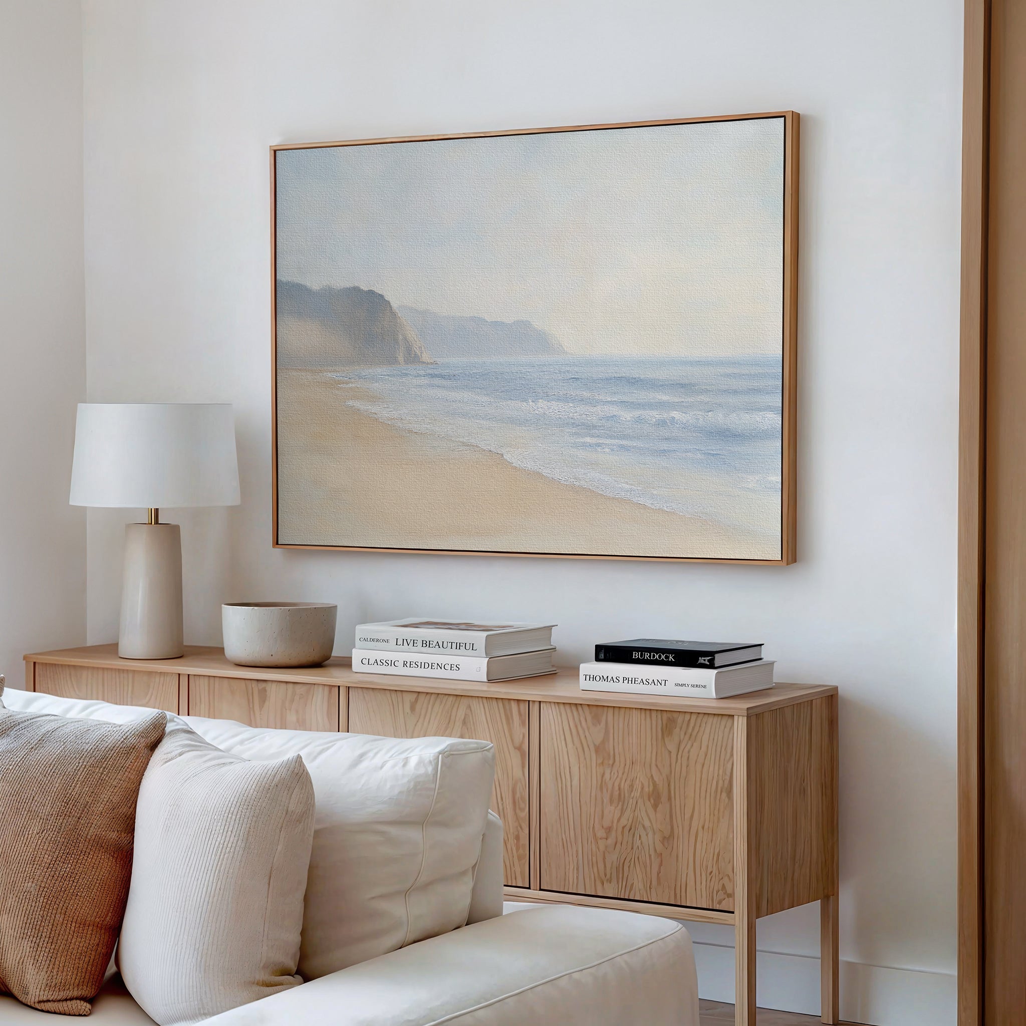 A minimalist living room features a light wood sideboard, stacked books, a white table lamp, a ceramic bowl, and the Coastal Cliffs Canvas Art—a large beach landscape—displayed above a cream sofa for modern decor flair.