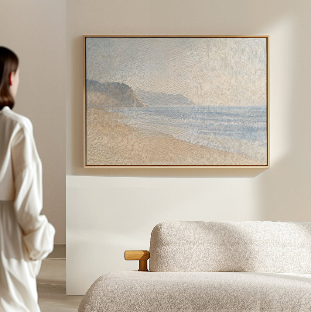 A woman in a white robe admires the Coastal Cliffs Canvas Art—a beach scene with soft waves—hanging above her cream sofa in a bright, minimal room, completing her modern home decor.