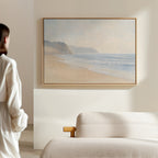 A woman in a white robe admires the Coastal Cliffs Canvas Art—a beach scene with soft waves—hanging above her cream sofa in a bright, minimal room, completing her modern home decor.
