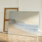 The Coastal Cliffs Canvas Art, featuring a calm beach and misty cliffs, leans against a wall on a wooden floor, perfect for modern decor with an additional blank canvas propped behind it.