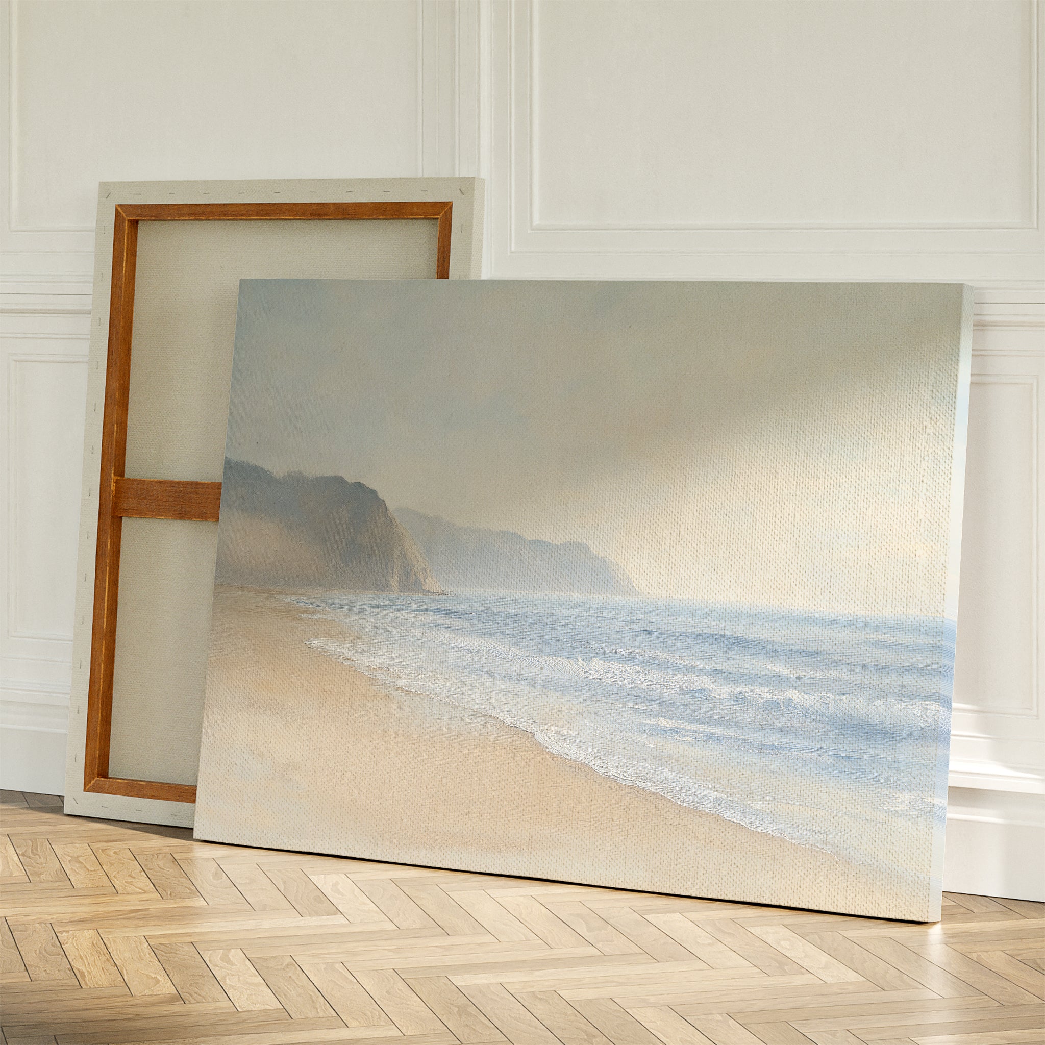 The Coastal Cliffs Canvas Art, featuring a calm beach and misty cliffs, leans against a wall on a wooden floor, perfect for modern decor with an additional blank canvas propped behind it.