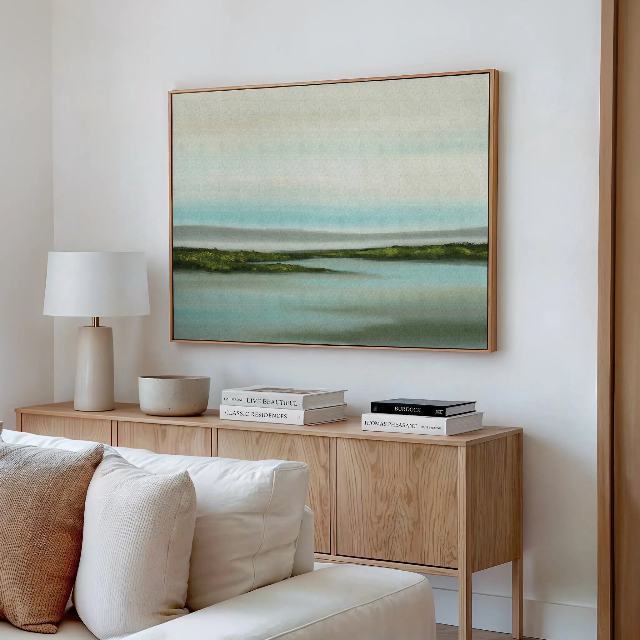 A modern living room with a light wood sideboard, white lamp, ceramic bowl, books, and the Coastal Marsh Canvas Art featuring blue and green tones above a white sofa.