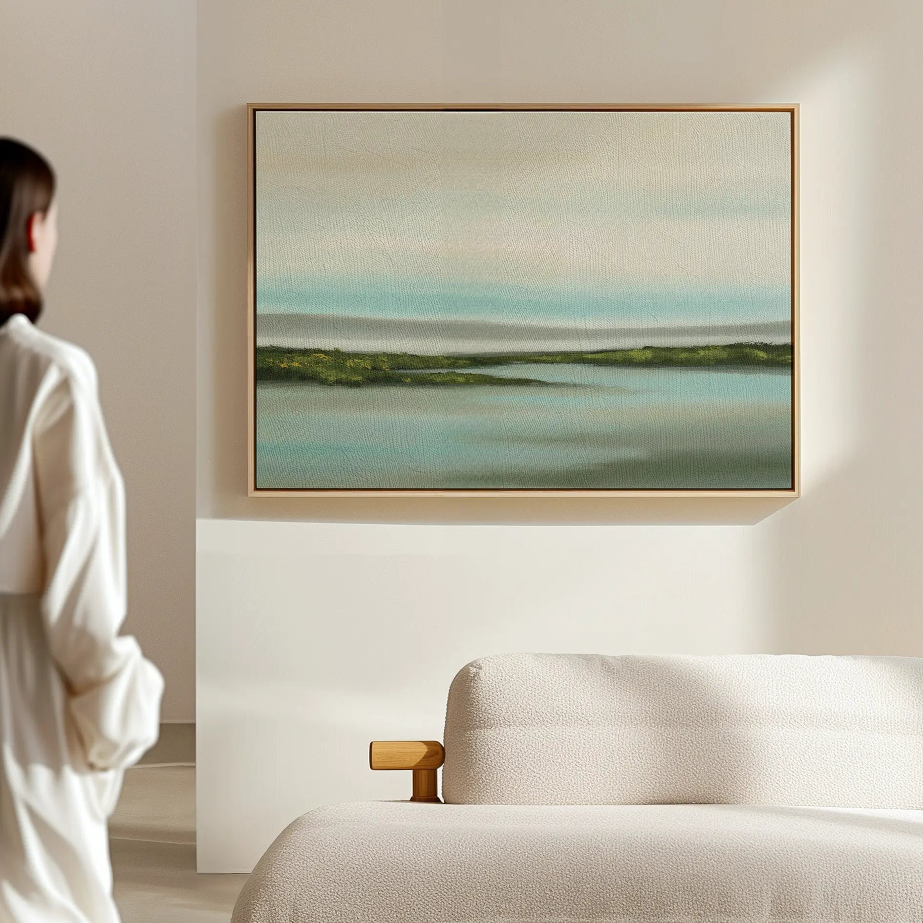 A person in a white robe stands in a minimalist room, gazing at the Coastal Marsh Canvas Art—a framed piece with soft blue, green, and beige tones—hanging above a light-colored sofa.
