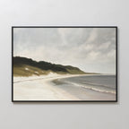 The Coastal Silence Canvas Art is a framed wall piece featuring a serene beach landscape with gentle waves, grassy dunes, and a cloudy sky, displayed on a light-colored wall.