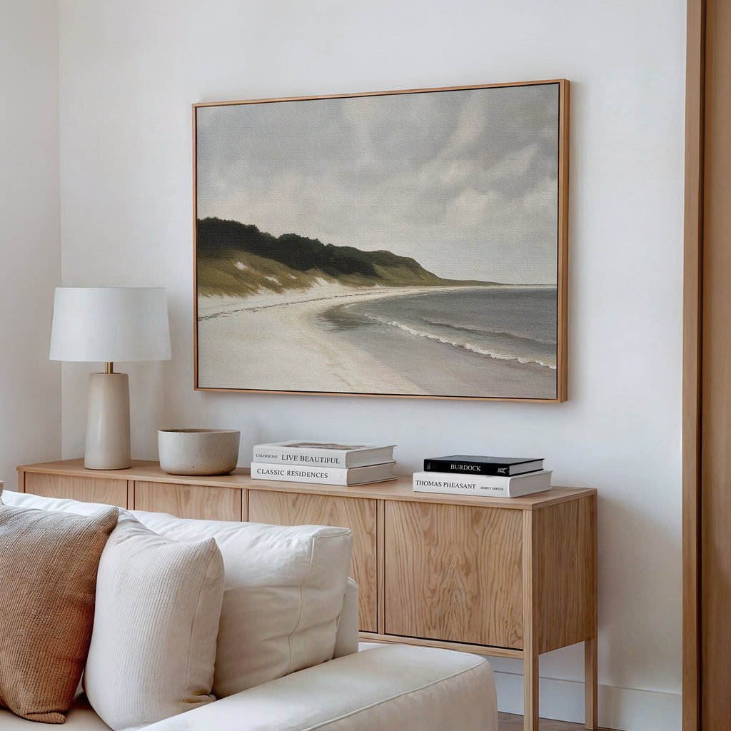 A modern living room with a light wood sideboard, stacked books, a lamp, a bowl, and the Coastal Silence Canvas Art hanging above a white sofa with cream cushions.