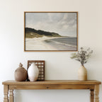 A wooden table with decorative vases and dried flowers stands against a white wall. Above it hangs the Coastal Silence Canvas Art, featuring a serene landscape of sandy beach, gentle waves, grassy dunes, and a cloudy sky.
