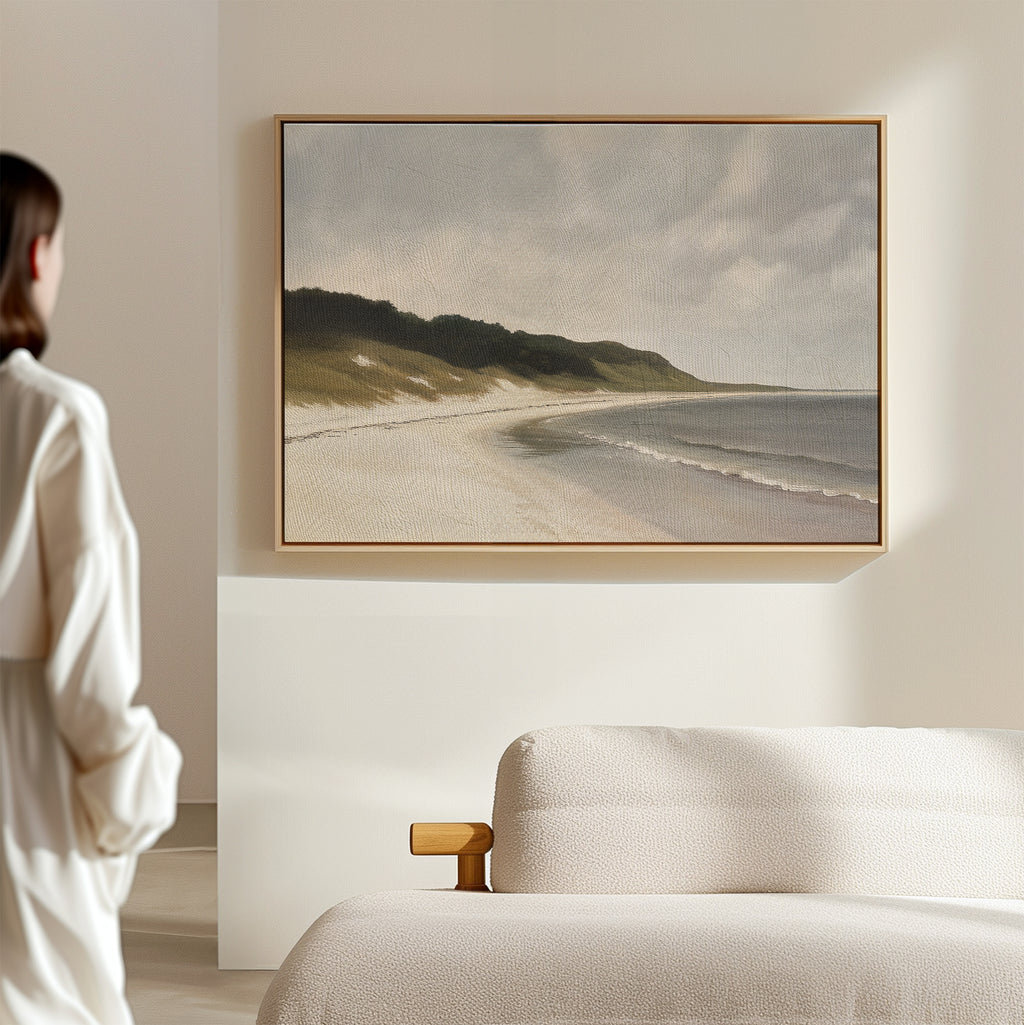 A person in a white outfit stands in a modern, minimal room with a light sofa, admiring the Coastal Silence Canvas Art featuring a tranquil beach and waves as serene coastal wall art.