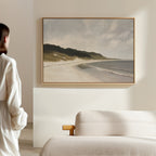 A person in a white outfit stands in a modern, minimal room with a light sofa, admiring the Coastal Silence Canvas Art featuring a tranquil beach and waves as serene coastal wall art.