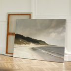 The Coastal Silence Canvas Art, featuring a sandy beach with gentle waves and distant greenery, leans against a white wall on a wooden floor with a blank canvas positioned behind it.
