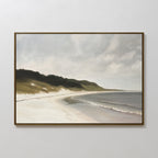 The Coastal Silence Canvas Art, featuring a quiet beach with gentle waves, sandy shore, and grassy dunes beneath a cloudy sky, hangs on a plain white wall.