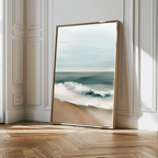 The Coastal Whisper Abstract Canvas Art, featuring ocean waves on a sandy beach, leans against an ornate white wall in a bright room with herringbone floors. Soft side light casts gentle shadows, making it stunning large wall art.