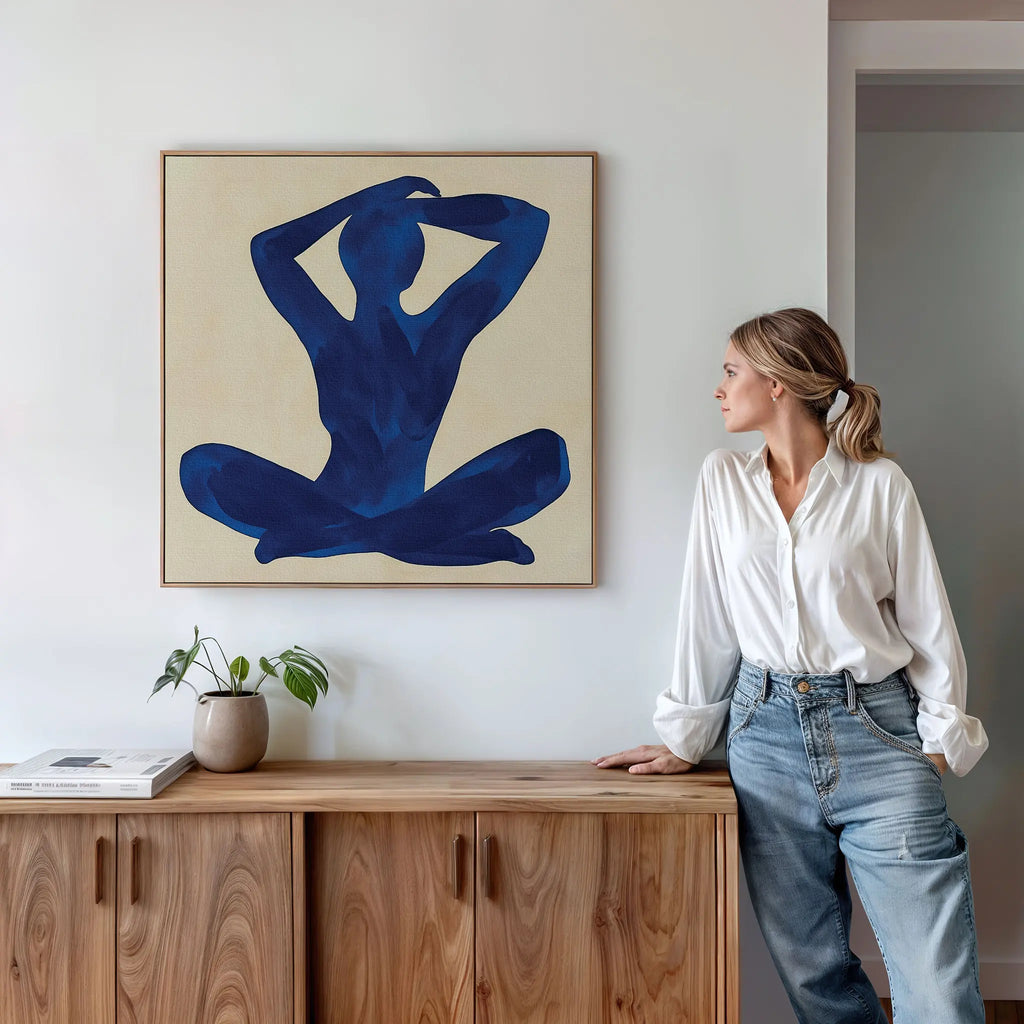 A woman in a white blouse and jeans leans against a wooden cabinet, gazing at the Cobalt Figure Canvas Art—a minimalist abstract blue figure—displayed on a white wall above her, with a small plant and books on the cabinet.