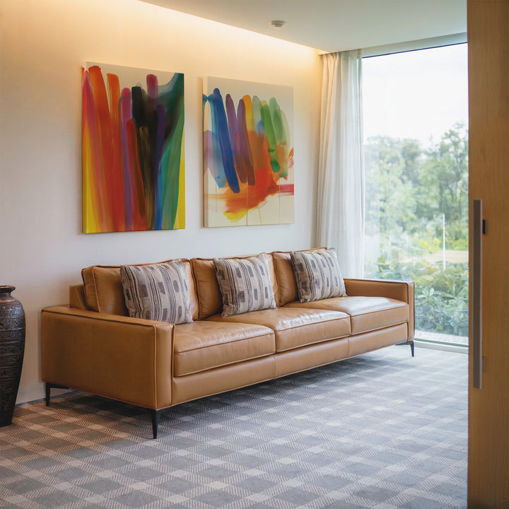 A modern living room with a tan leather sofa, three patterned pillows, the Color Cascade Set of 2 as vibrant abstract wall art, a large window with sheer curtains, and a decorative vase on the floor.