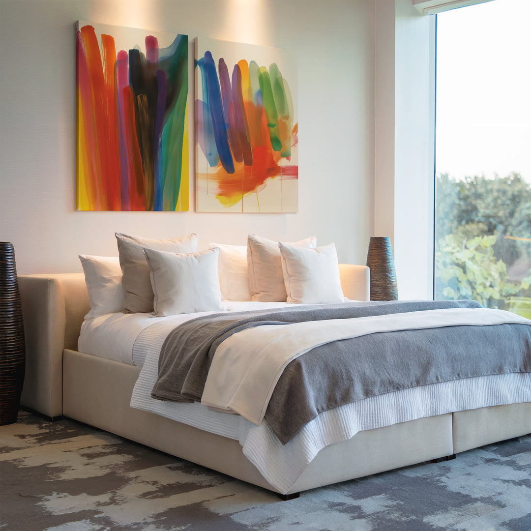 A modern bedroom features a large bed with white bedding and a gray blanket. Above the bed hangs the Color Cascade Set of 2 abstract prints, while two tall, dark vases on either side complement the bright, airy space.