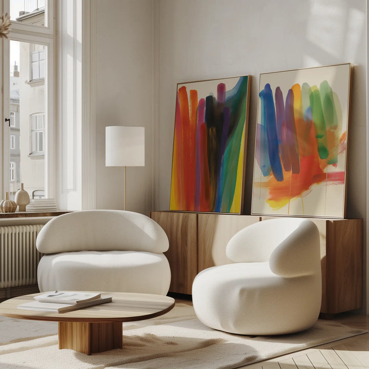 A modern living room with two white curved chairs, a round wooden coffee table, and a wooden sideboard. The Color Cascade Set of 2 abstract wall art rests on the sideboard beneath tall windows, adding vibrant flair to the decor.