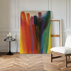 The Color Cascade Abstract Canvas Art, featuring bold vertical brushstrokes, leans against a white wall in a bright room with an empty wooden frame, modern side table with candles, and a cushioned chair on herringbone floors.