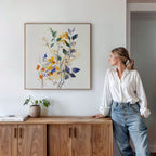A woman in a white blouse and jeans leans against a wooden sideboard, admiring the Colorful Branches Canvas Art above. A potted plant and books sit nearby in a softly lit, minimalist living room.