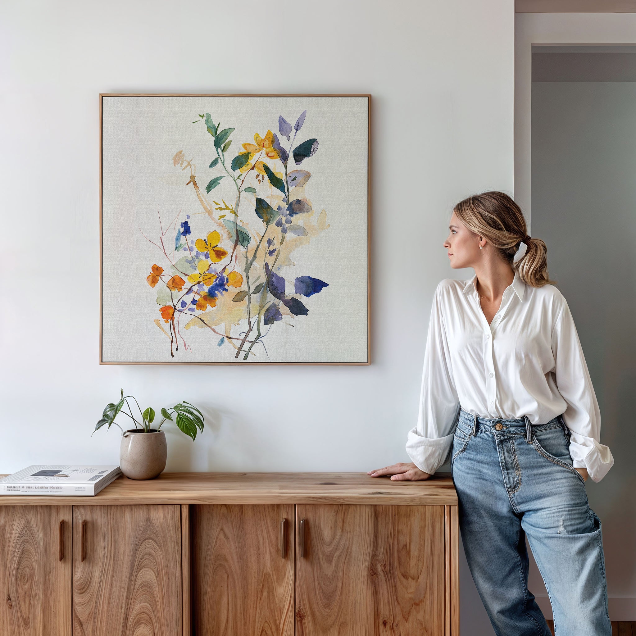 A woman in a white blouse and jeans leans against a wooden sideboard, admiring the Colorful Branches Canvas Art above. A potted plant and books sit nearby in a softly lit, minimalist living room.