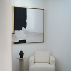 A modern, minimalist corner features a cream armchair, a small pedestal with a black vase, a white accent lamp, and the Monochrome Harmony Abstract Canvas Art in a gold frame for an elevated touch of modern wall decor.