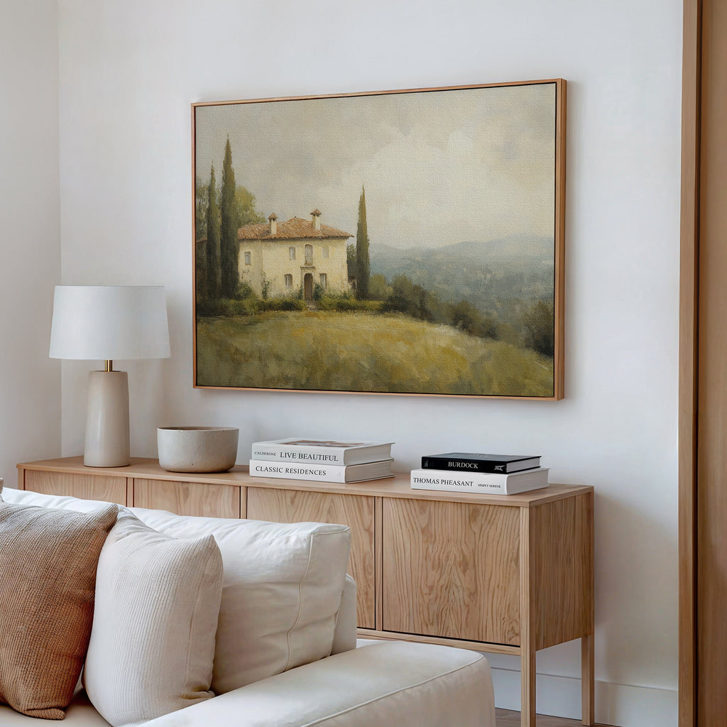 A cozy living room features a light wood console topped with books, a lamp, and a bowl. Above, the Country Villa Canvas Art adds charm to the neutral decor, creating an inviting atmosphere.