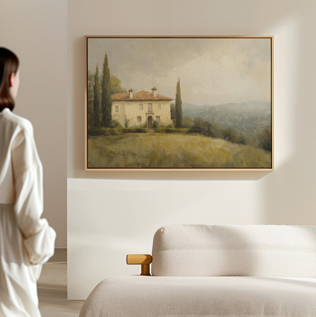 A person in a white robe admires the Country Villa Canvas Art—rustic wall decor of a house amid trees and grassy hills—displayed above a cream sofa in a minimal, light-filled living room with serene beige walls.