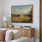 A cozy living room with a light wood sideboard, neutral decor, and a white table lamp. Above the sideboard hangs the Countryside Dream Canvas Art, depicting a manor house in a green field with cows and distant hills.