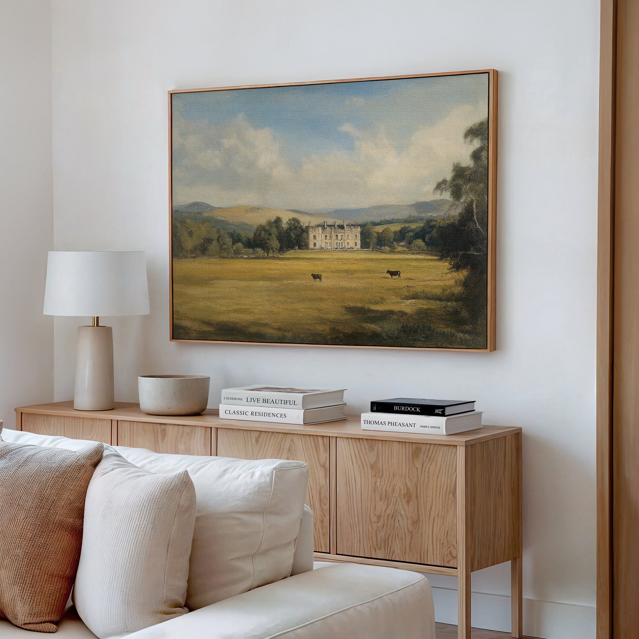 A cozy living room with a light wood sideboard, neutral decor, and a white table lamp. Above the sideboard hangs the Countryside Dream Canvas Art, depicting a manor house in a green field with cows and distant hills.