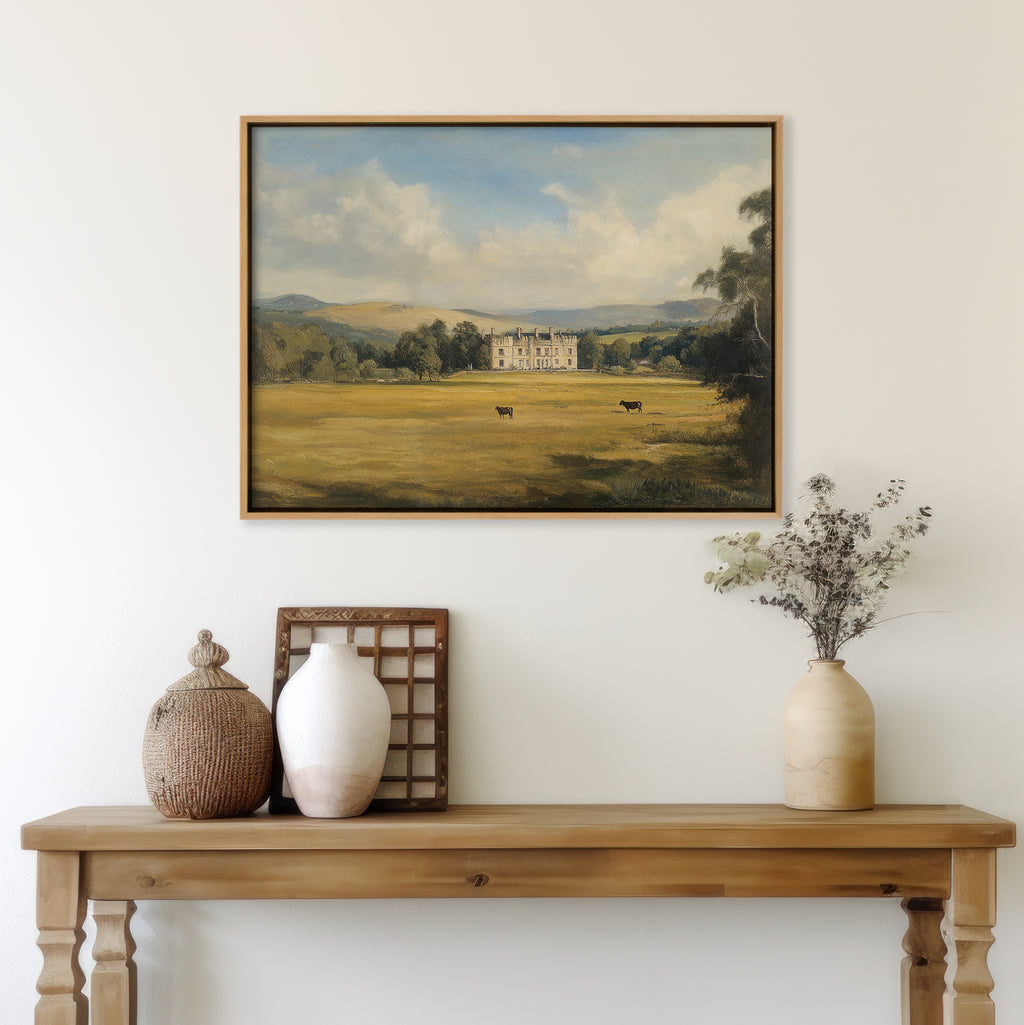 A wooden table with ceramic vases, a wicker jar, dried flowers, and rustic decor sits below the Countryside Dream Canvas Art—a framed landscape of a manor and cows—hung on a white wall above.