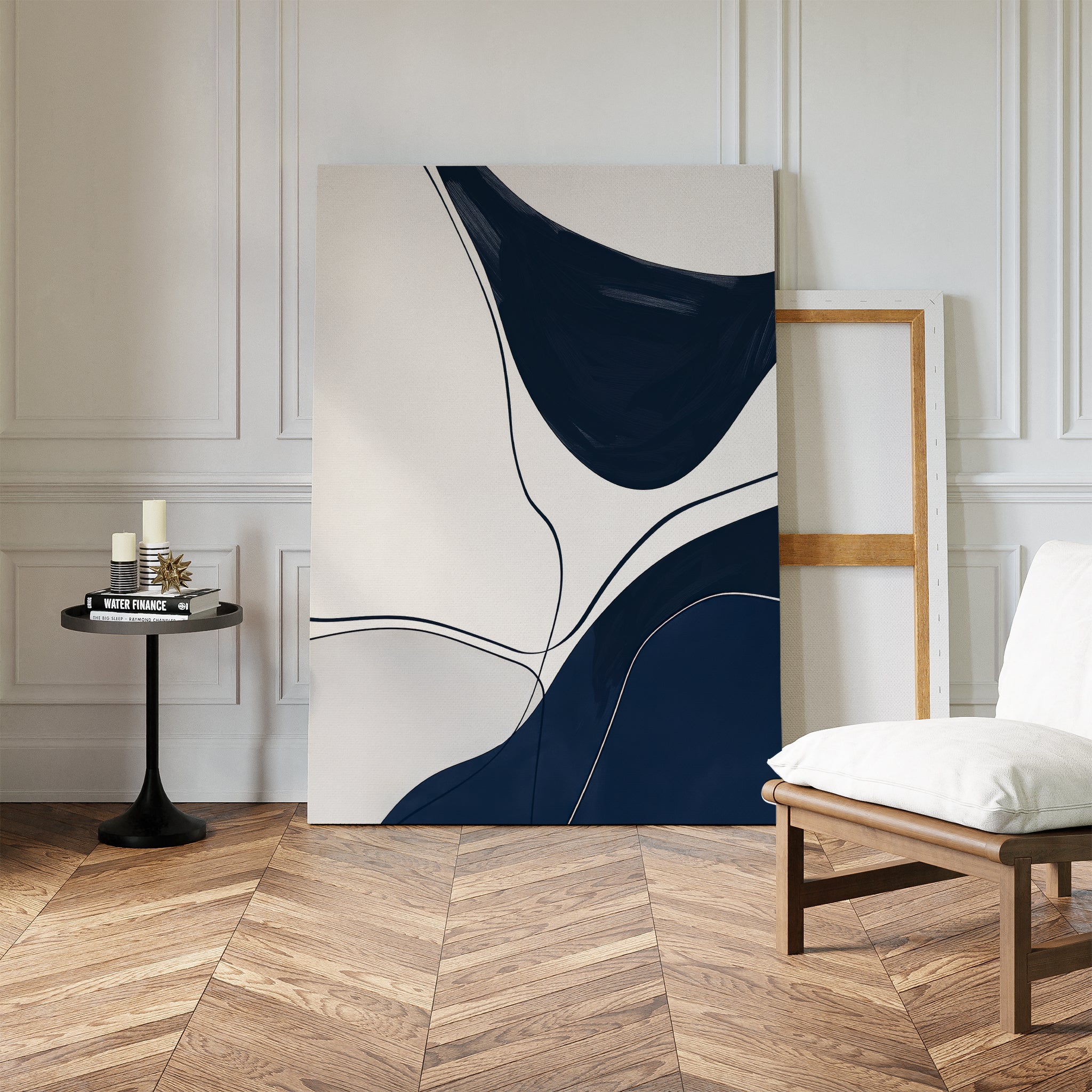 The Curved Lines Abstract Canvas Art, featuring navy blue and cream curves, leans against a white wall beside minimalist decor: a wooden frame, cushioned white chair, and round black table with candles on herringbone floors.