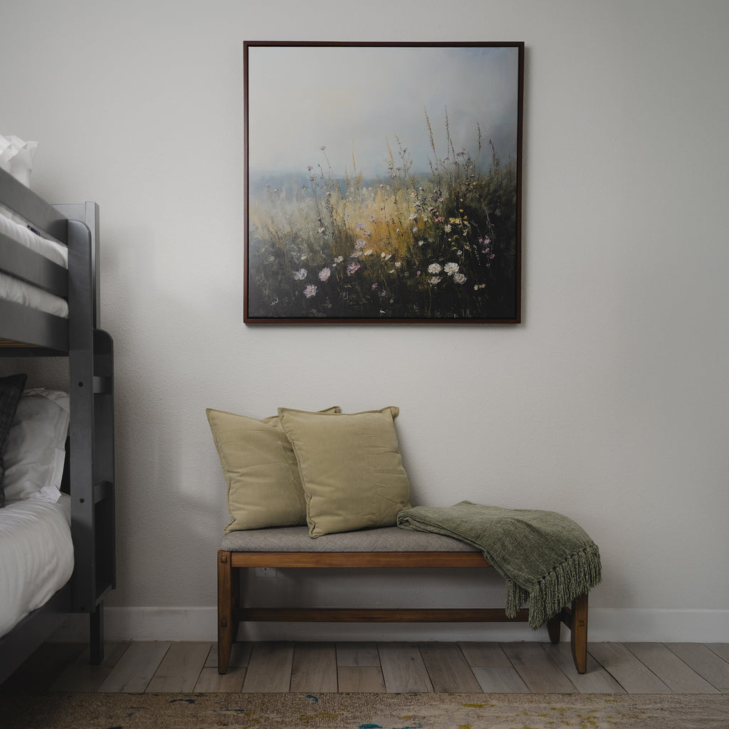 A wooden bench with two beige cushions and a green throw blanket sits against a white wall, beneath Wildflowers Canvas Art. Bunk beds are partially visible on the left, on a light wood floor.