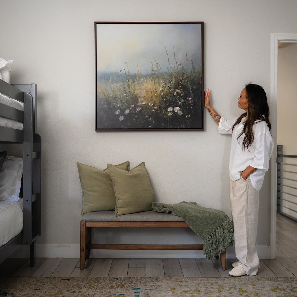 A woman in light clothes admires Wildflowers Canvas Art above a bench with sage green pillows and throw, next to a bunk bed—a charming addition to living room decor.