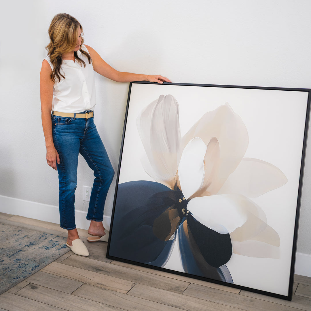 A woman in a white top and jeans stands indoors, holding the Whispering Petals Canvas Art—minimalist wall decor with abstract floral shapes in neutral and dark tones, perfect for enhancing your living room style.