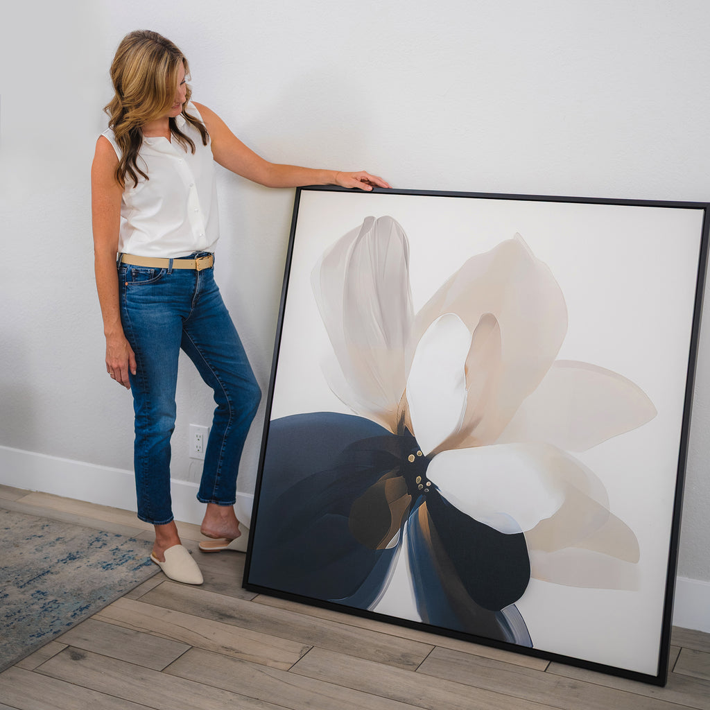 A woman in a white sleeveless top and jeans stands indoors, leaning the Whispering Petals Canvas Art—featuring soft neutral and dark petals—against a white wall, making it perfect for effortless living room decor.