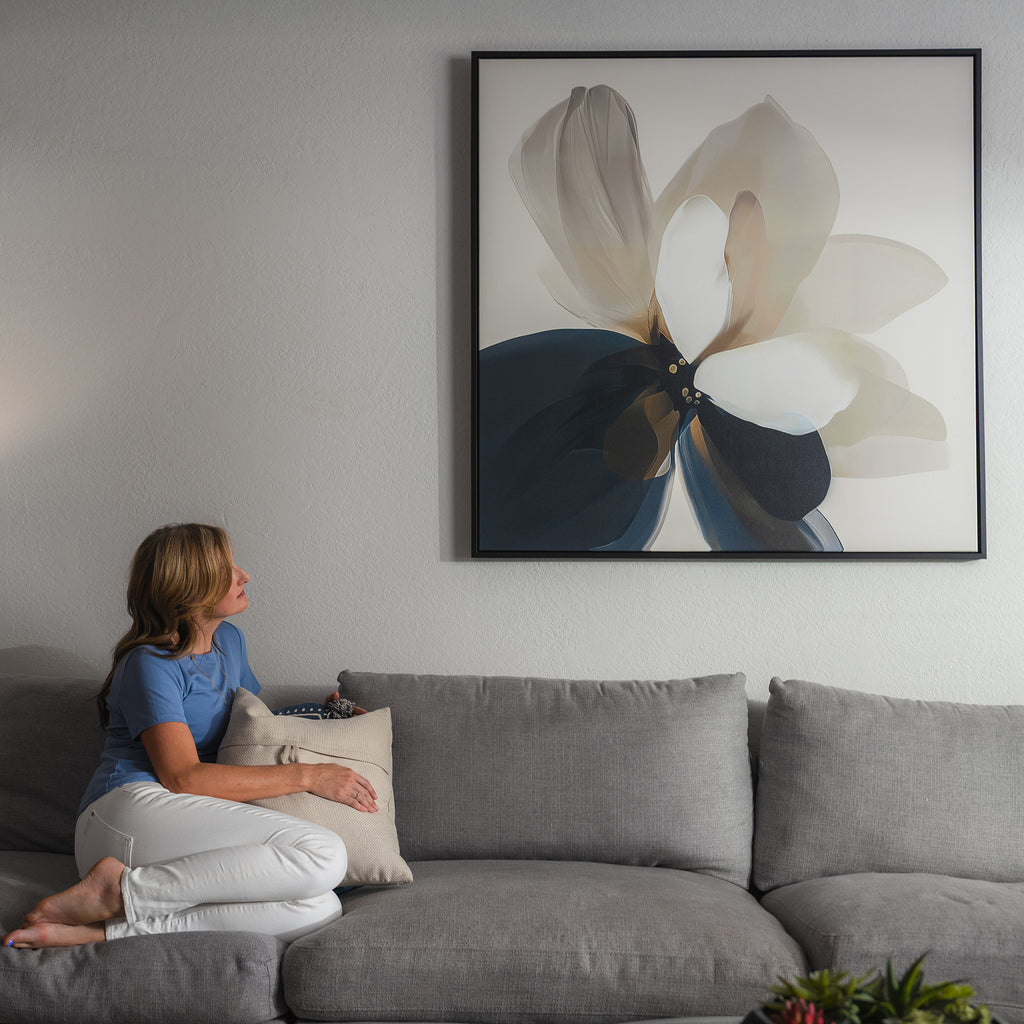 A woman sits on a gray couch, holding a pillow and admiring the Whispering Petals Canvas Art—a large abstract flower painting in neutral tones—enhancing the living room decor above her.
