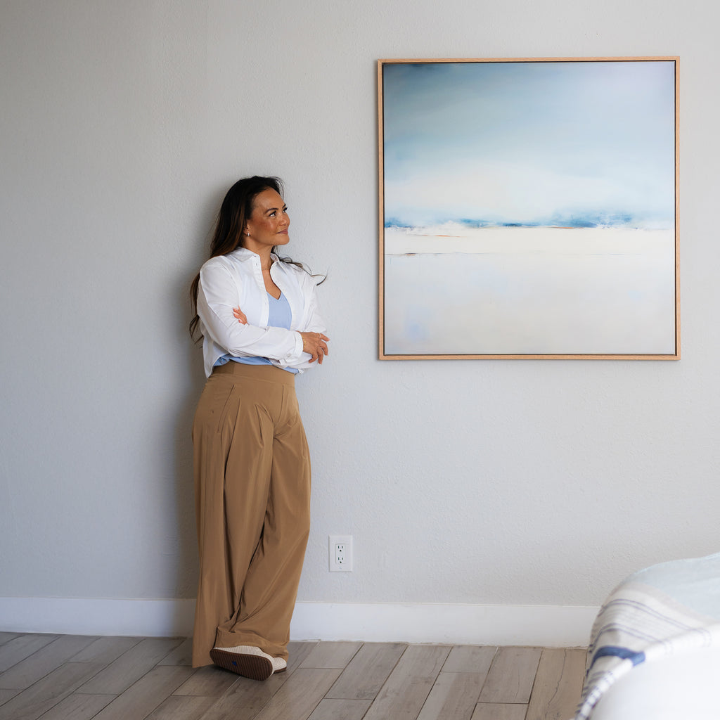 A woman in a white blouse and tan pants stands with arms crossed, smiling in a bright room. Beside her hangs the Misty Morning Abstract Canvas Art, its blue and white tones enhancing the tranquil atmosphere.
