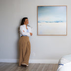 A woman in a white blouse and tan pants stands with arms crossed, smiling in a bright room. Beside her hangs the Misty Morning Abstract Canvas Art, its blue and white tones enhancing the tranquil atmosphere.