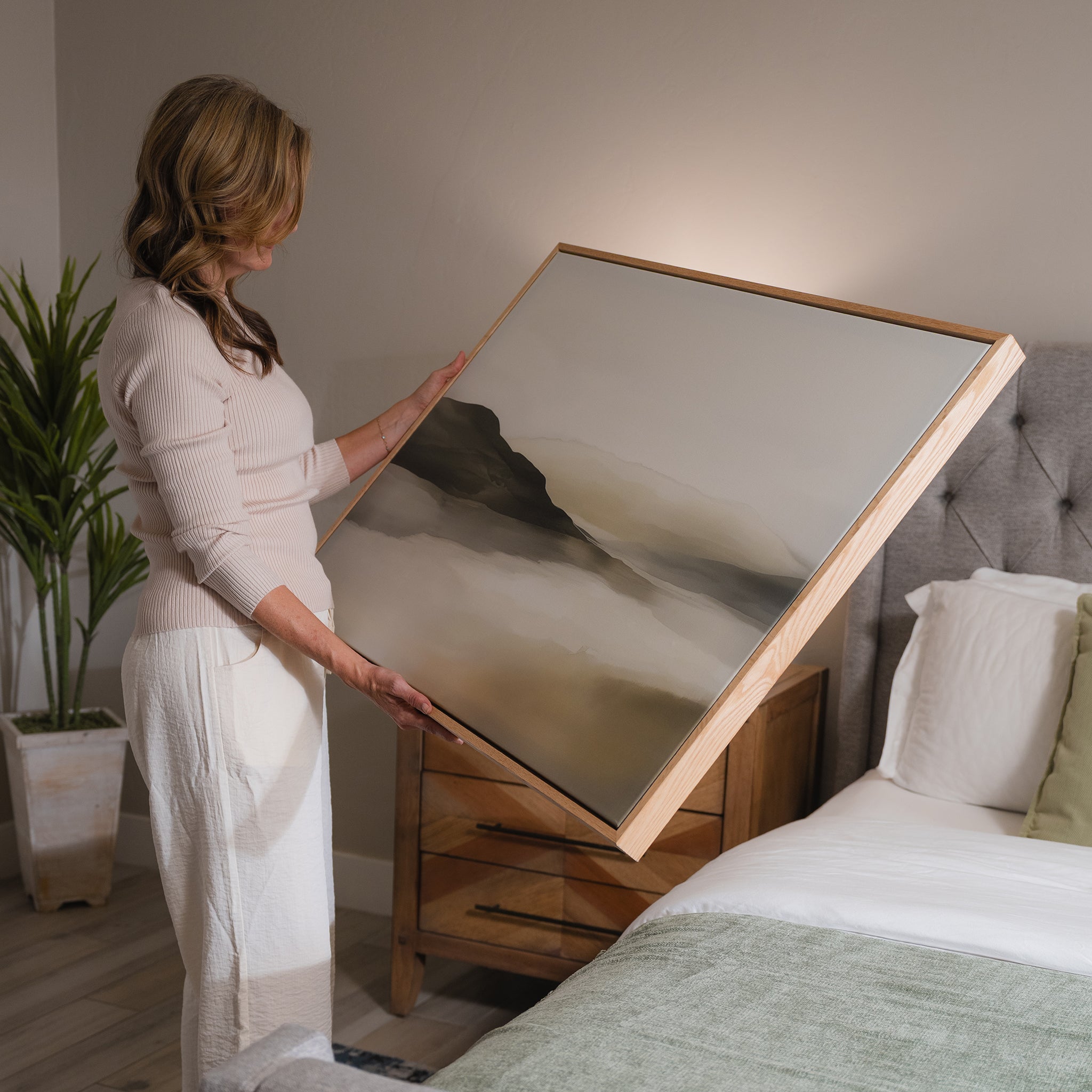 A woman stands beside a bed, holding the Soft Valley Canvas Art – Horizontal, featuring soft, earthy tones. The minimalist room includes a potted plant, wooden nightstand, and upholstered headboard.