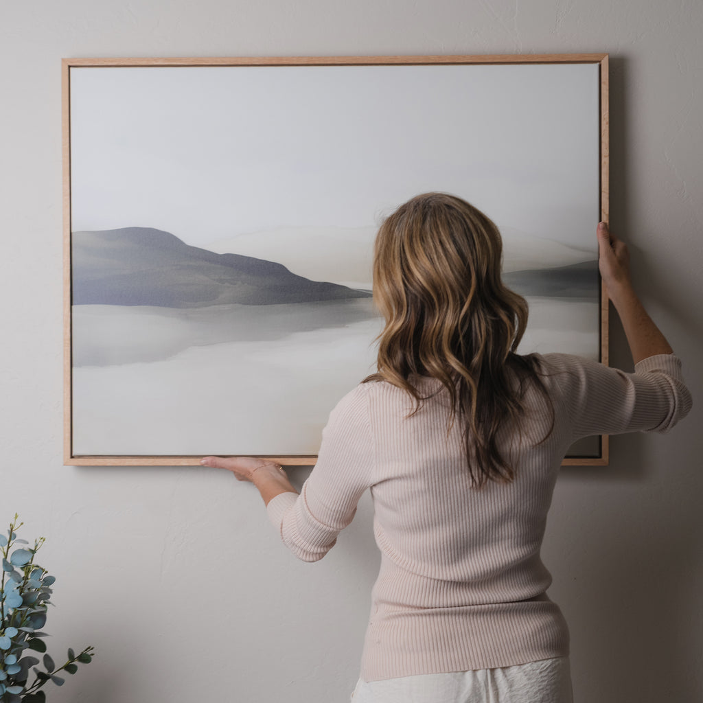 A woman with wavy hair hangs the Soft Valley Canvas Art – Horizontal, featuring a misty mountain landscape, on a light-colored wall. A small leafy plant in the corner enhances the minimalist decor.