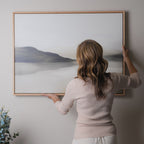 A woman with wavy hair hangs the Soft Valley Canvas Art – Horizontal, featuring a misty mountain landscape, on a light-colored wall. A small leafy plant in the corner enhances the minimalist decor.