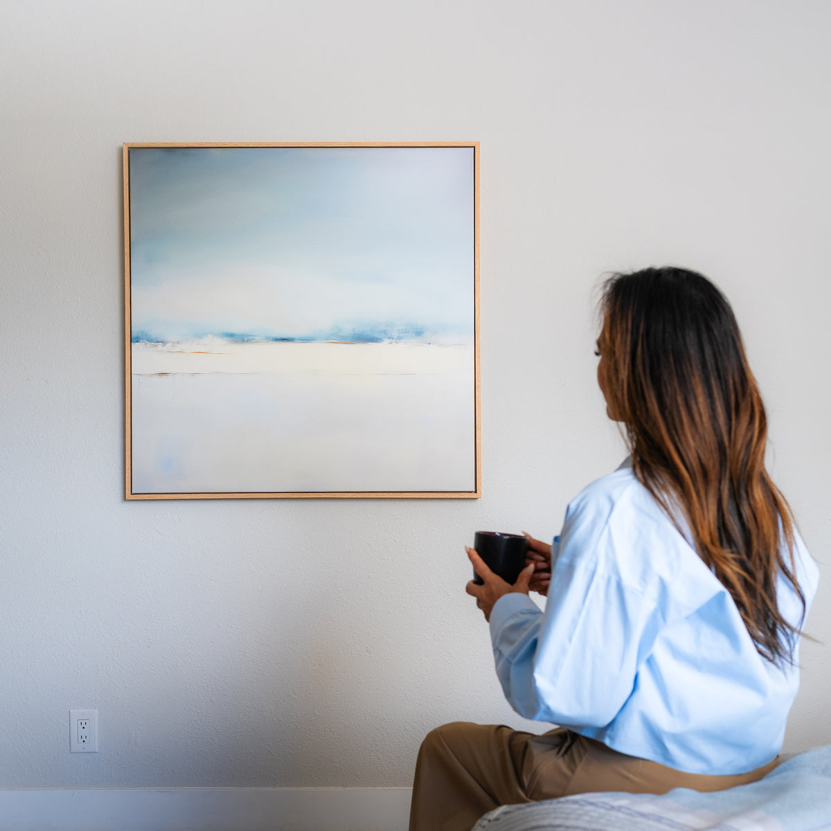 A person with long hair sits on a bed holding a mug, gazing at the Misty Morning Abstract Canvas Art in soft blue and white tones on a plain wall, creating a tranquil atmosphere.