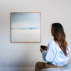 A person with long hair sits on a bed holding a mug, gazing at the Misty Morning Abstract Canvas Art in soft blue and white tones on a plain wall, creating a tranquil atmosphere.