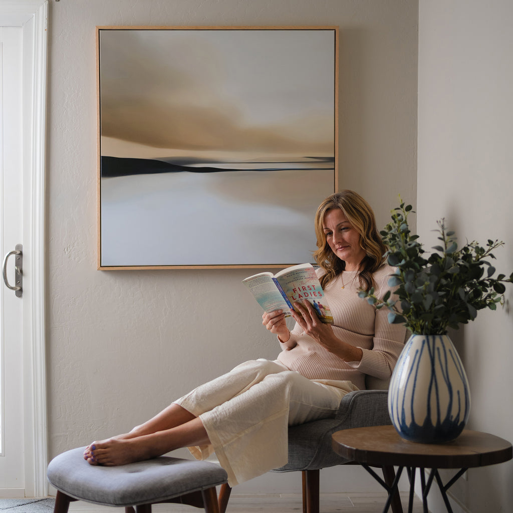 A woman relaxes on a chair with her feet on an ottoman, reading in a modern living room. Above her hangs the Desert Quietude Abstract Canvas Art, adding style to the decor, while a vase with greenery decorates a small table nearby.
